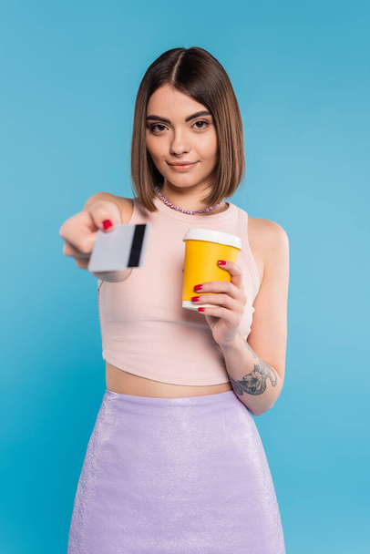 to go coffee, happy young woman with short hair, tattoos and nose piercing holding paper cup and giving credit card on blue background, generation z, summer trends, attractive, coffee culture  - Photo, Image
