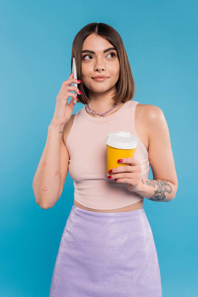 to go coffee, happy and tattooed young woman with short hair and nose piercing holding paper cup and talking on smartphone on blue background, generation z, summer trends, attractive, coffee culture  - Photo, Image