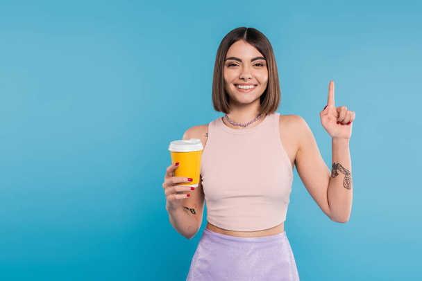 to go coffee, happy young woman with short hair, tattoos and nose piercing holding paper cup on blue background, generation z, summer trends, attractive, coffee culture, everyday style, pointing up - Photo, Image