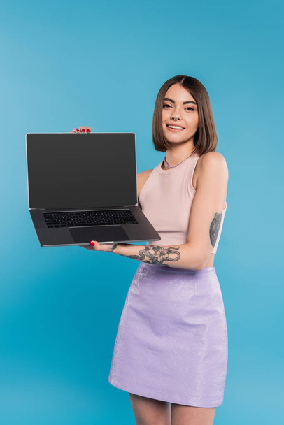 freelancer, cheerful young woman with short hair, tattoos and nose piercing holding laptop on blue background, generation z, summer trends, attractive, remote work, everyday style, blank screen - Photo, Image