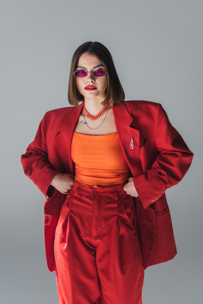 professional attire, young brunette woman with short hair posing in pink sunglasses and red suit on grey background, generation z, trendy outfit, fashionable model, full length  - Fotografie, Obrázek