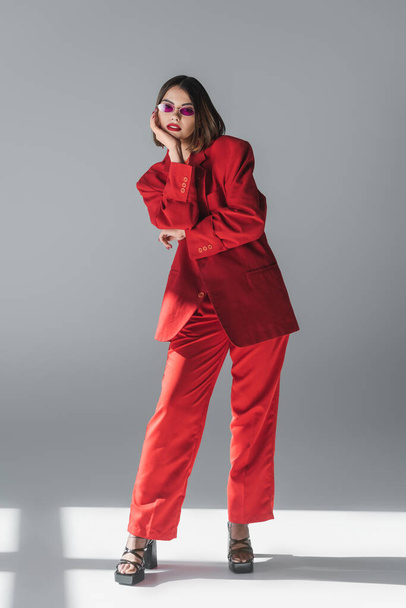 lady in red, young brunette woman with short hair posing in pink sunglasses and red suit on grey background, generation z, trendy outfit, fashionable model, executive style  - Φωτογραφία, εικόνα
