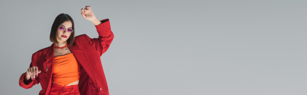 gen z à la mode, jeune femme brune aux cheveux courts posant en lunettes de soleil roses et costume rouge sur fond gris, génération z, tenue tendance, mannequin, style exécutif, bannière - Photo, image