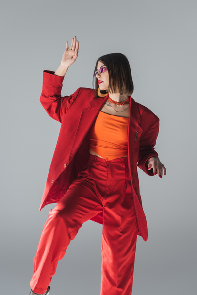 lady in red, young brunette woman with short hair posing in pink sunglasses and red suit and gesturing on grey background, generation z, trendy outfit, fashionable model, professional attire  - Photo, Image