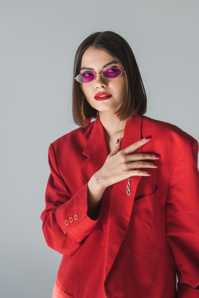 contemporary fashion, young brunette woman with short hair posing in pink sunglasses and red suit on grey background, generation z, trendy outfit, fashionable model, professional attire  - Fotografie, Obrázek