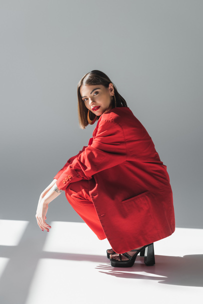 full length, lady in red, young brunette woman with short hair sitting in suit on grey background, generation z, trendy outfit, fashionable model, professional attire, executive style, heeled shoes - 写真・画像