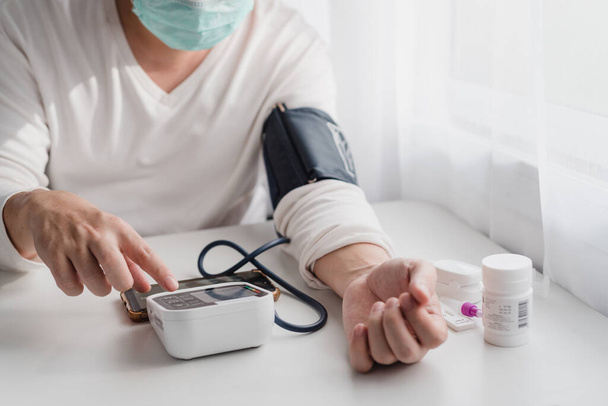 顔のマスクを身に着けている人は、デジタル血圧計と薬のボトルをテーブルの上に使用しています,在宅医療の概念. - 写真・画像