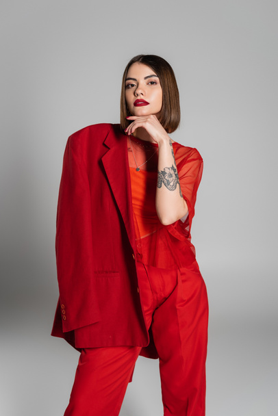 trendy outfit, bold makeup, tattooed woman with short hair holding red blazer on grey background, generation z, professional attire, executive style, young professional  - Foto, immagini