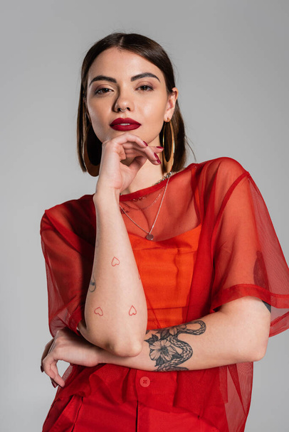 fashion look, red outfit, tattooed and brunette woman with short hair and nose piercing posing in transparent blouse on grey background, modern style, generation z, portrait  - Fotografie, Obrázek