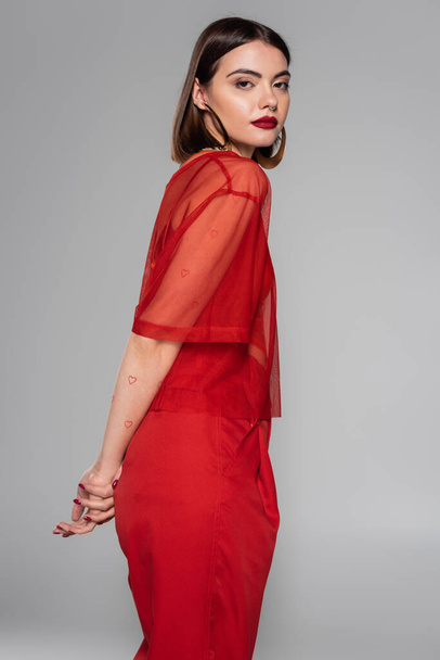 stylish red outfit, hoop earrings, tattooed and brunette woman with short hair and nose piercing posing in transparent blouse and pants on grey background, modern style, generation z, fashion - Photo, Image