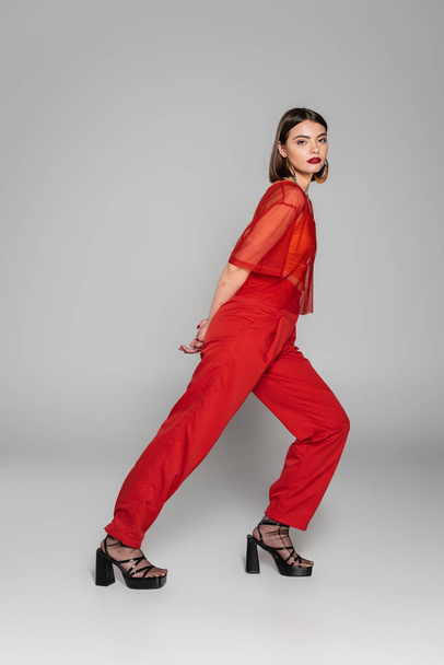 model in red outfit, tattooed and brunette woman with short hair and nose piercing posing in transparent blouse and pants on grey background, modern style, generation z, fashion trend, full length  - Foto, Imagem