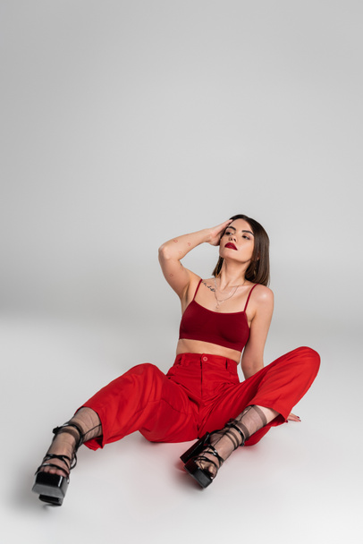 fashion trend, chic style, young model in red outfit, tattooed woman with short hair and nose piercing posing in red crop top and pants on grey background, generation z, full length  - Photo, Image