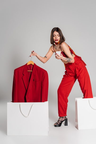 consumerism, tattooed woman with short hair and nose piercing holding hanger with blazer and using smartphone near shopping bags on grey background, modern fashion trend, full length  - 写真・画像