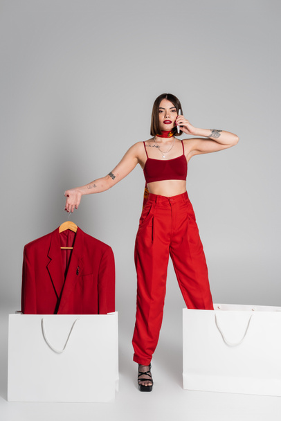 consumerism, tattooed woman with brunette short hair and nose piercing holding hanger with blazer and talking on smartphone near shopping bags on grey background, modern fashion trend, full length  - 写真・画像