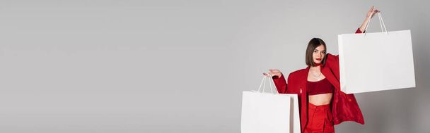 consumismo, jovem mulher com cabelo curto morena e nariz piercing segurando sacos de compras e andando em fundo cinza, tendência de moda moderna, roupa na moda, terno vermelho, banner  - Foto, Imagem