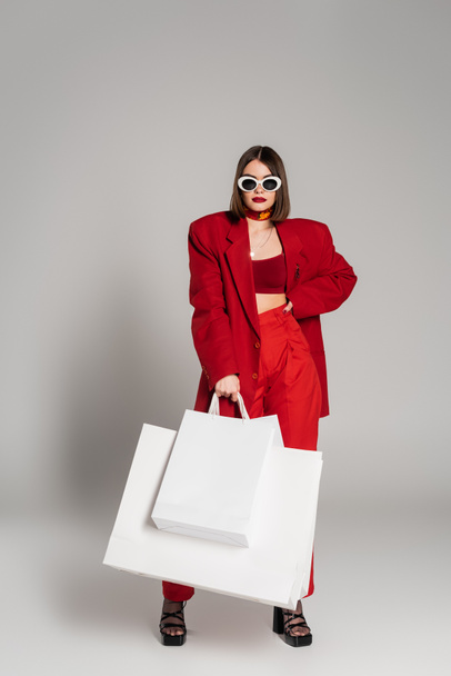 red suit, generation z, young woman with short hair and nose piercing posing in sunglasses and holding shopping bags on grey background, youth culture, consumerism  - Foto, imagen