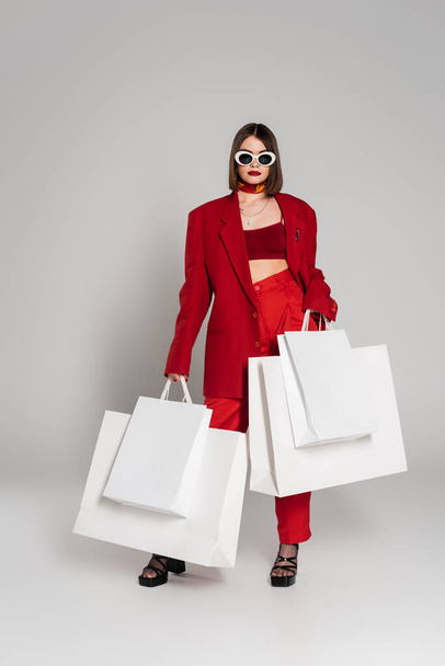 generation z, young woman with brunette short hair and nose piercing posing in sunglasses and red suit while holding shopping bags on grey background, modern fashion, consumerism, full length   - Fotoğraf, Görsel