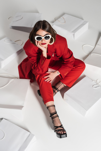 consumismo, mujer joven con pelo corto morena, piercing en la nariz y tatuaje posando en gafas de sol y traje rojo mientras se sienta alrededor de bolsas de compras sobre fondo gris, vista de ángulo alto  - Foto, imagen