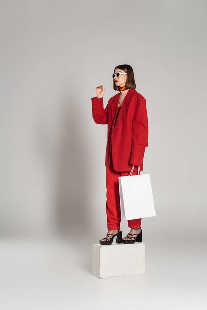Generation z, Konsum, junge Frau mit brünetten kurzen Haaren und Nasenpiercing posiert in Sonnenbrille und rotem Anzug, während sie Einkaufstasche in der Hand hält und auf Betonwürfel auf grauem Hintergrund steht  - Foto, Bild