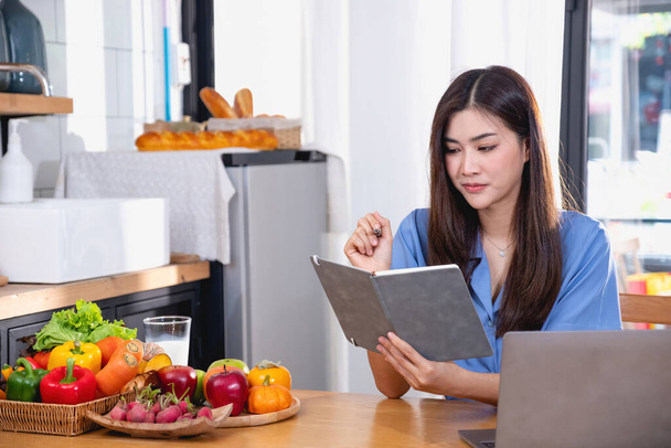 Una mujer joven con una hermosa cara en una camisa azul con el pelo largo comiendo fruta sentada dentro de la cocina en casa con un ordenador portátil y portátil para la relajación, Concept Vacation.  - Foto, imagen