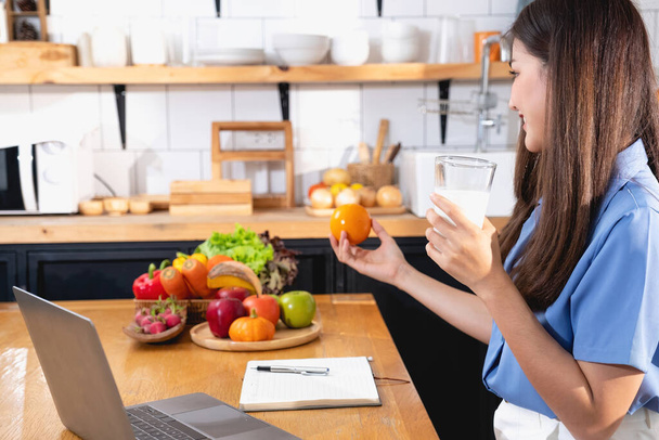 Een jonge vrouw met een mooi gezicht in een blauw shirt met lang haar eten fruit zitten in de keuken thuis met een laptop en notebook voor ontspanning, Concept vakantie.  - Foto, afbeelding