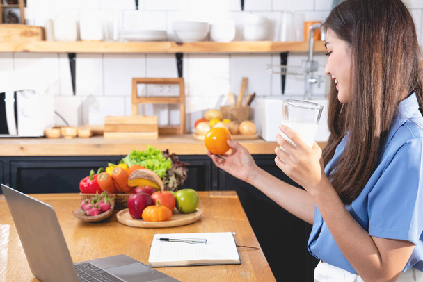 Eine junge Frau mit einem schönen Gesicht in einem blauen Hemd mit langen Haaren, die zu Hause in der Küche sitzt und mit Laptop und Notizbuch zur Entspannung Obst isst, Concept Vacation.  - Foto, Bild