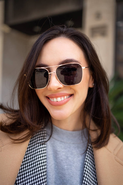 Streetstyle, street fashion concept: woman wearing trendy outfit walking in city. Cream trench coat, sunglasses. Looking camera, vertical photo close up - Foto, Imagem