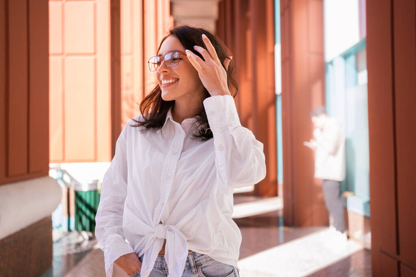 Successful smiling business woman wearing eyeglasses correct hairstyle by hand. Modern businesswoman smile standing outdoor dressed white shirt looking away. Business woman happy positive emotions - Фото, зображення