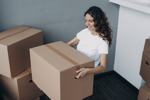 Smiling hispanic female homeowner with packed box, moving into new apartment. Happy girl settling in rented house. - Foto, Imagem