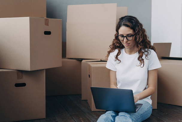 Focused woman in glasses reviews moving arrangements online, sitting amidst cardboard boxes, searching for a reliable mover. - Zdjęcie, obraz