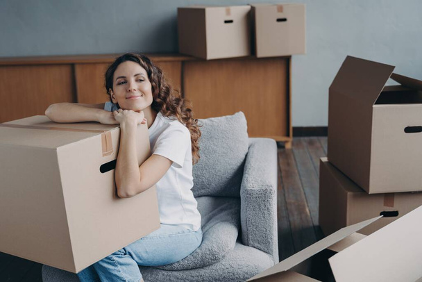 Happy homeowner resting on armchair with a cardboard box during relocation. Spanish woman relaxing after packing, awaiting transportation for moving to new house. - Photo, Image