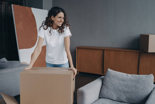 Happy young Spanish girl in new home apartment with a cardboard box. Pleased woman prepares for house renovation or relocation, dreaming of modern interior design. - Photo, Image