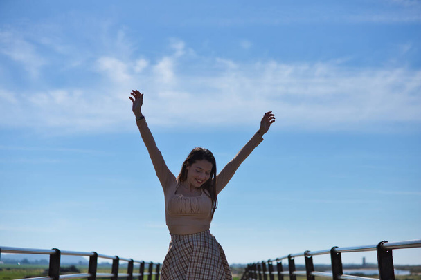 Mooie jonge vrouw met lang bruin haar draagt een kort rokje en shirt heft haar armen vrolijk op als ze de houten brug oversteekt. Op de achtergrond de blauwe lucht en witte wolken. - Foto, afbeelding