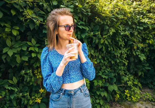 Beautiful woman in blue blouse and sunglasses in green park smiling drinking iced coffee and enjoying summer sunshine. Coffee break on work. - 写真・画像