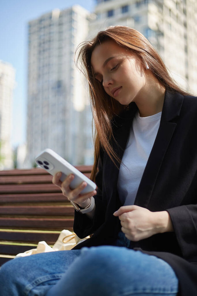 美しいブルネットの女性は、ワイヤレスヘッドフォンと現代的なスマートフォンでオンラインで音楽を聴く。ベンチに座って携帯電話を使っている若い女性の肖像画 - 写真・画像