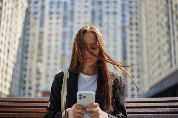 Beautiful young woman with long brown hair typing a message on a smart phone outdoor in windy sunny day. Portrait of a pretty white female person with a hair covering her face in wind - Фото, изображение