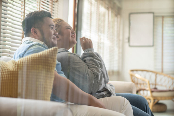 Young Asian chubby couple watching tv series and movie on the couch in living room. Man and woman enjoying a fun time together at home. People laughing and smiling together - Photo, image
