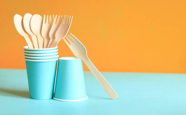 Wooden disposable forks and spoons in a blue paper cup, side view. Eco friendly disposable kitchen utensils on orange background, copy space. Ecological items concept - Photo, Image