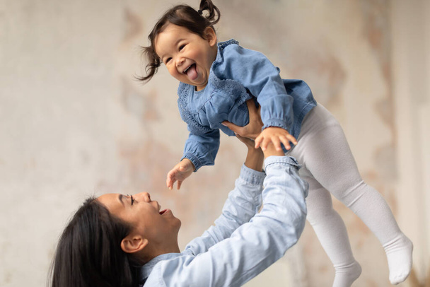 Vidám ázsiai anya játszik a baba lánya, felemel és lovagol csecsemő karokban, nevetve megosztása örömteli pillanatokat otthon. Mami ölelkezik és élvezi, hogy gondoskodhat az imádnivaló gyerekéről. - Fotó, kép