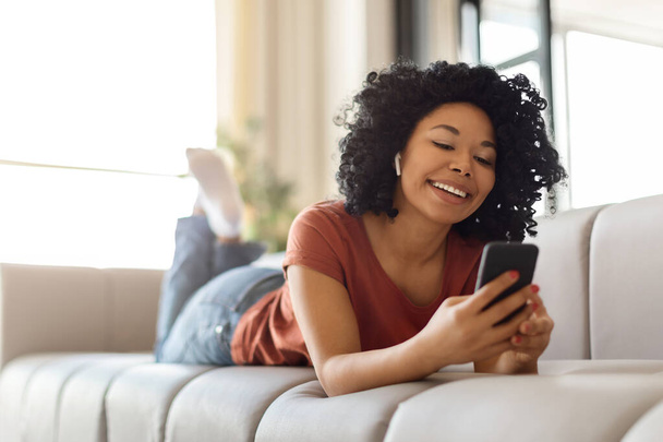 Porträt einer lächelnden jungen schwarzen Frau mit drahtlosen Ohrhörern, die sich zu Hause mit dem Smartphone entspannt, einer glücklichen Afroamerikanerin, die ihr Handy benutzt, während sie auf der Couch liegt und Lieblingsmusik genießt - Foto, Bild