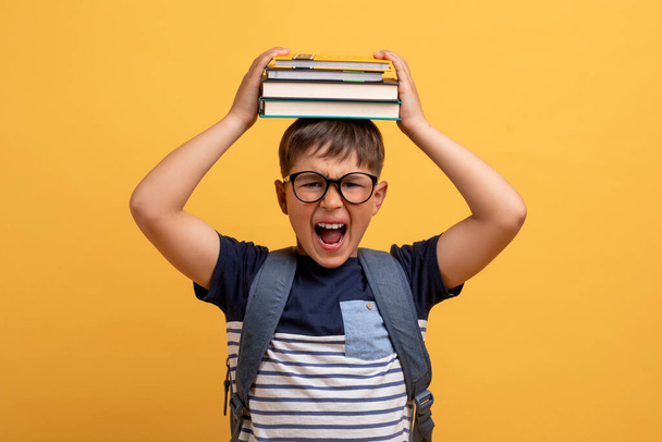 バックパックを持って眼鏡をかけ、彼の頭の上に本を身に着けているかわいい男の子の学校を強調悲鳴,学校での困難の苦しみ,黄色のスタジオの背景に隔離 - 写真・画像