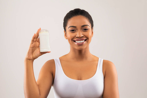 Vidám, fiatal afro-amerikai nő fehér felsőben, kezében dezodorral, szürke stúdió háttéren mosolyogva, másolja a helyet. Testápolás, hónalj, hónalj higiénia - Fotó, kép