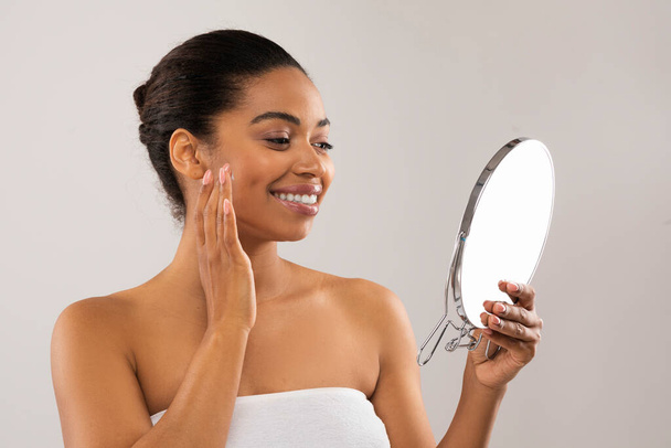 Rotina de cuidados com a pele. Feliz atraente mulher millennial afro-americana olhando para o pequeno espelho, massageando rosto com os dedos e sorrindo, aplicando creme, soro ou óleo, isolado em cinza, espaço de cópia - Foto, Imagem