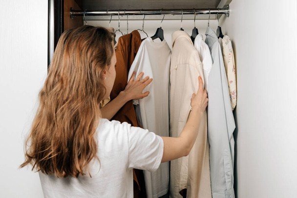 A young Caucasian woman chooses shirts and t-shirts from a wardrobe hanging on hangers in a closet. The concept of changing the wardrobe from summer to winter, buying new clothes - Photo, Image