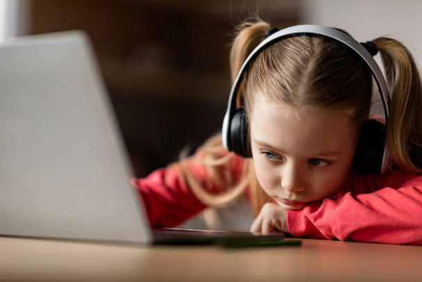 Depressed Little Girl Looking At Laptop Screen And Leaning On Desk At Home, Closeup Portrait Of Preteen Female Kid Wearing Wireless Headphones Feeling Bored, Tired Of E-Learning And Online Study - Zdjęcie, obraz