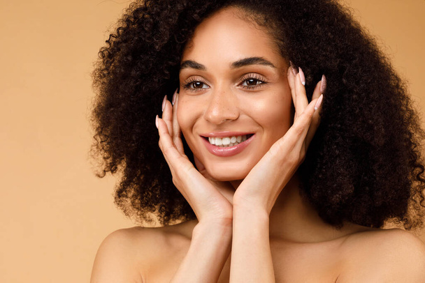 Headshot of happy young black lady touching her cheeks, profiter d'une peau soyeuse après avoir utilisé des produits de soins du visage de beauté premium bio de luxe, fond beige, gros plan - Photo, image