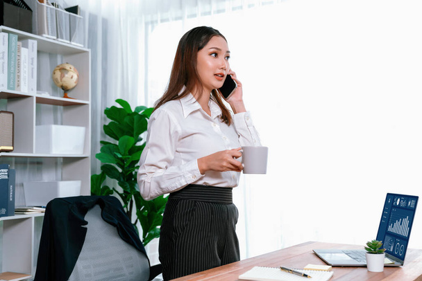 Young enthusiastic businesswoman talking on business call and writing notes on her laptop as multitask office lady. Female employee writing business tasks while talking on phone call with clients. - Foto, Imagen