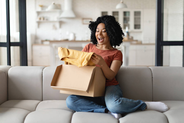 Glückliche junge schwarze Frau packt Paket mit neuen Kleidern zu Hause aus, freudige Afroamerikanerin öffnet Karton und nimmt stilvolles Hemd heraus, genießt Shopping und Online-Einkäufe - Foto, Bild