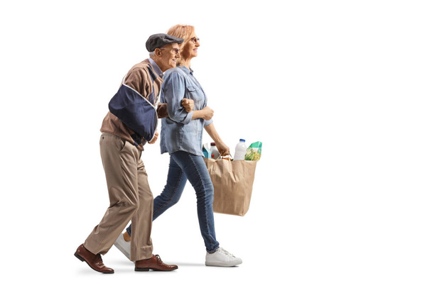 Full length profile shot of a woman helping an elderly man with an injured arm and carrying grocery bags isolated on white background - Photo, Image