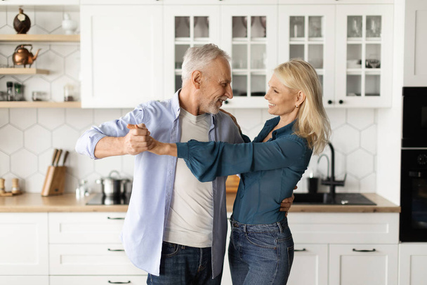Portret van romantisch senior paar dansen samen in keuken interieur, gelukkig volwassen echtgenoten die handen vasthouden en kijken naar elkaar, genieten van tijd doorbrengen met elkaar thuis, kopiëren ruimte - Foto, afbeelding